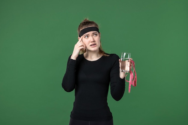 Vista frontal de las mujeres jóvenes con un vaso de agua y medir la cintura sobre fondo verde, cuerpo deportivo, salud, entrenamiento negro, color, atleta