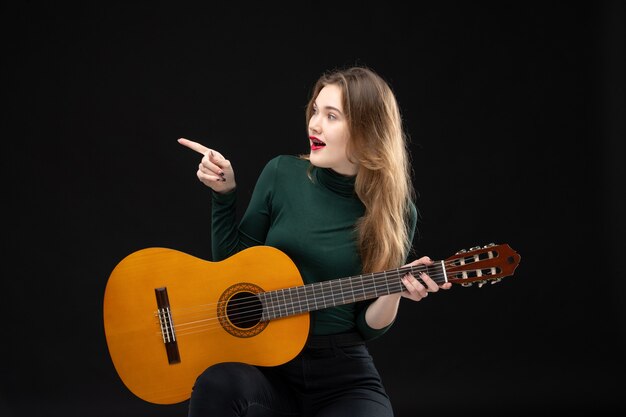 Vista frontal del músico femenino feliz sosteniendo la guitarra y apuntando algo en el lado derecho en negro