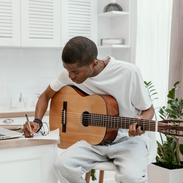Vista frontal del músico masculino en casa tocando la guitarra y escribiendo letras