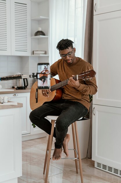 Vista frontal del músico masculino en casa tocando la guitarra y grabando con el teléfono inteligente