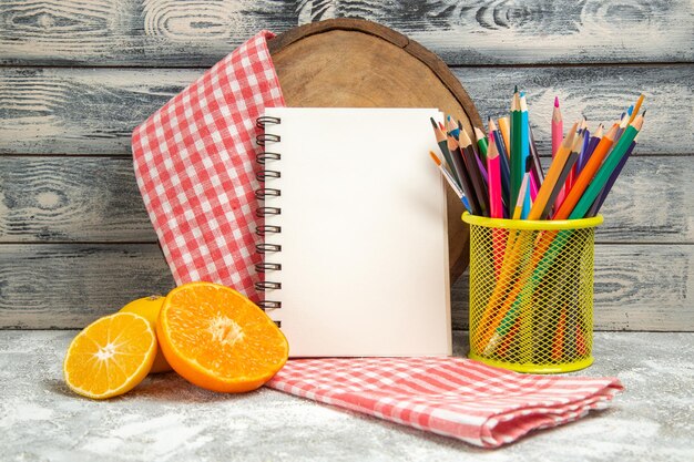 Vista frontal de naranjas en rodajas frescas con bloc de notas y lápiz sobre fondo gris color de cuaderno de frutas cítricas