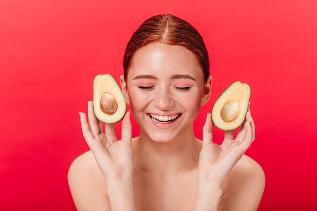 Vista frontal de la niña con aguacate sonriendo sobre fondo rojo. Foto de estudio de mujer dichosa riendo con los ojos cerrados.