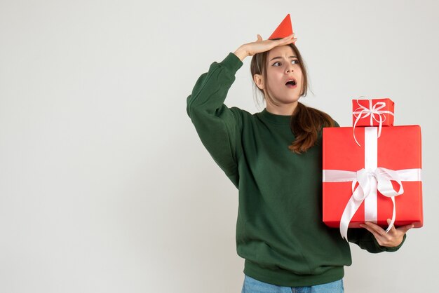 Vista frontal niña curiosa con gorro de fiesta observando sosteniendo sus regalos de navidad