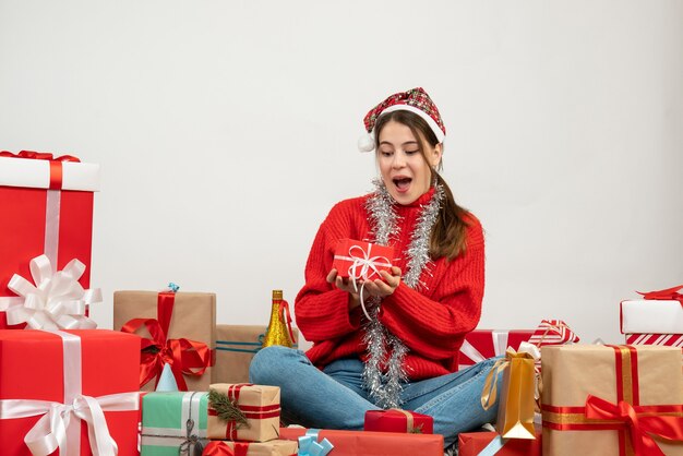 Vista frontal niña dichosa con sombrero de santa sosteniendo presente sentado alrededor de regalos