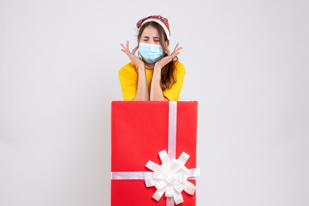 Vista frontal niña enojada con gorro de Papá Noel cerrando sus oídos con los dedos detrás de un gran regalo de Navidad