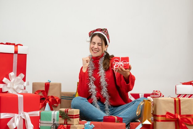 Vista frontal niña eufórica con sombrero de santa sosteniendo su regalo mientras se regocija sentado alrededor de regalos