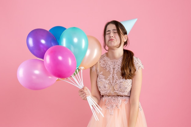 Vista frontal niña llorando con gorro de fiesta sosteniendo globos