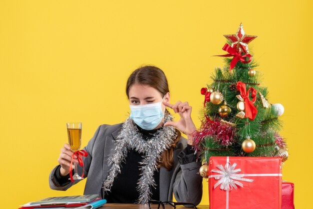 Vista frontal de la niña con máscara médica sentada en la mesa parpadeando el ojo proponiendo un brindis por el árbol de Navidad y regalos cóctel