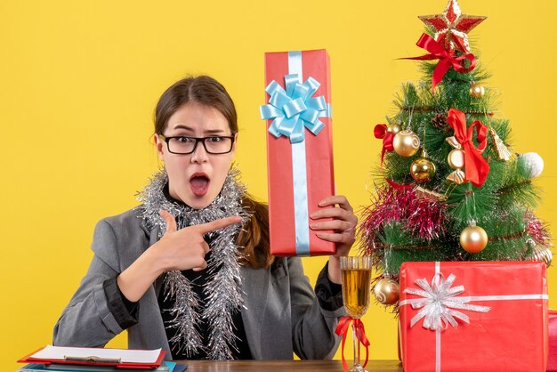 Vista frontal de la niña de ojos abiertos con anteojos sentado en la mesa mostrando regalo árbol de Navidad y cóctel de regalos