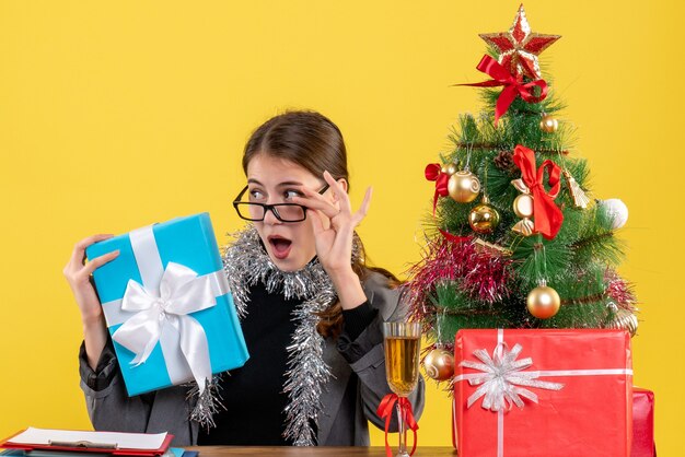 Vista frontal de la niña de ojos abiertos con anteojos sentado en la mesa quitándose los anteojos árbol de Navidad y cóctel de regalos