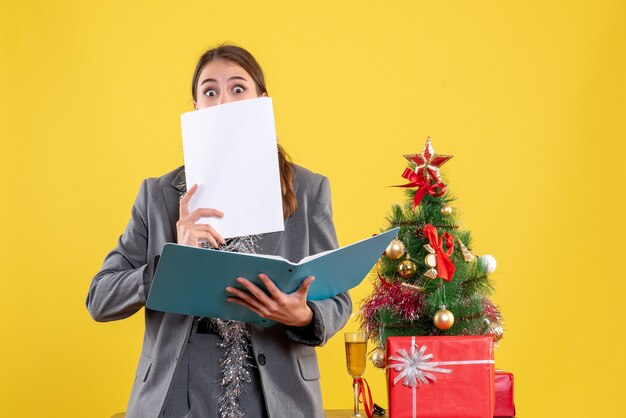 Vista frontal de la niña con los ojos abiertos comprobando documentos de pie cerca del árbol de Navidad y cóctel de regalos