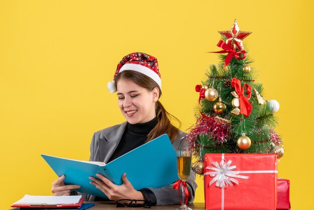 Vista frontal de la niña sonriente con sombrero de Navidad sentado en la mesa mirando documentos en la carpeta azul árbol de Navidad y cóctel de regalos