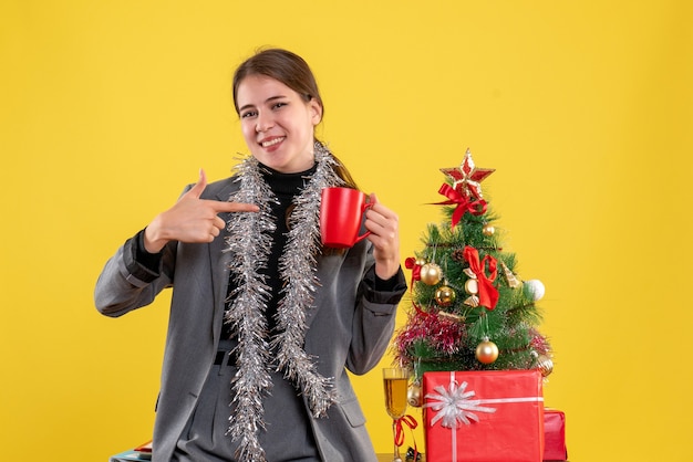 Vista frontal de la niña sonriente sosteniendo la taza roja apuntando con el dedo cerca del árbol de Navidad y cóctel de regalos