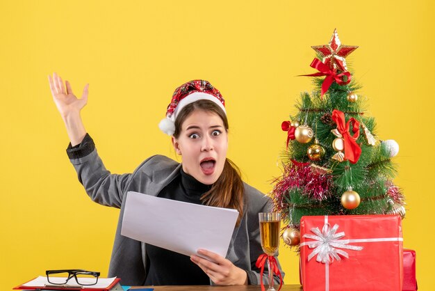 Vista frontal niña sorprendida con sombrero de Navidad sentado en la mesa árbol de Navidad y cóctel de regalos