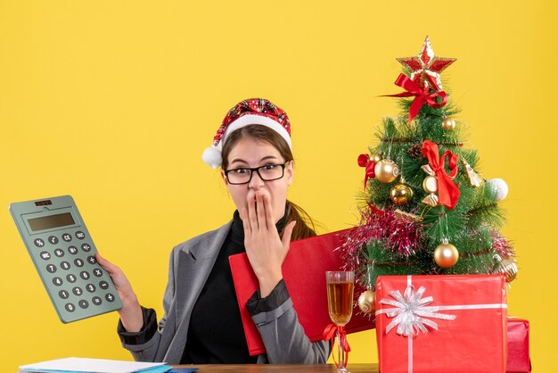 Vista frontal niña sorprendida con sombrero de Navidad sentado en la mesa mirando calculadora poniendo mano