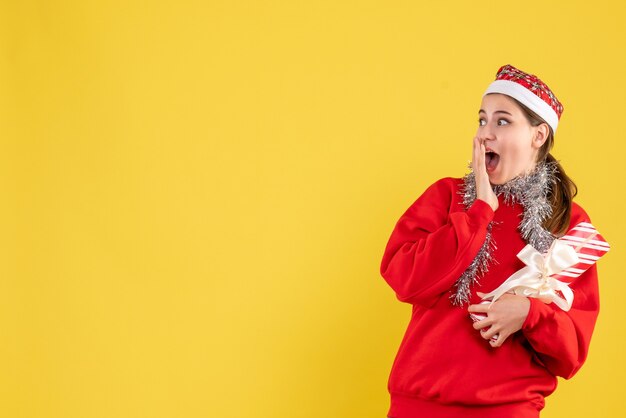 Vista frontal niña sorprendida con suéter rojo y gorro de Papá Noel sosteniendo su regalo