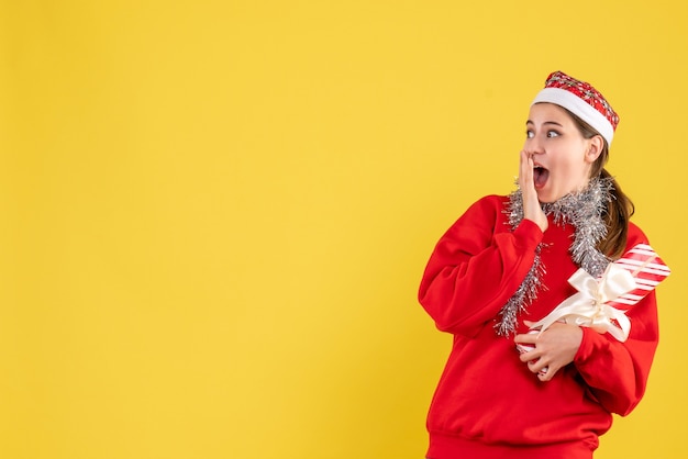 Vista frontal niña sorprendida con suéter rojo y gorro de Papá Noel sosteniendo su regalo