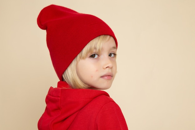 Vista frontal niño lindo en camisa roja y gorra en la pared blanca