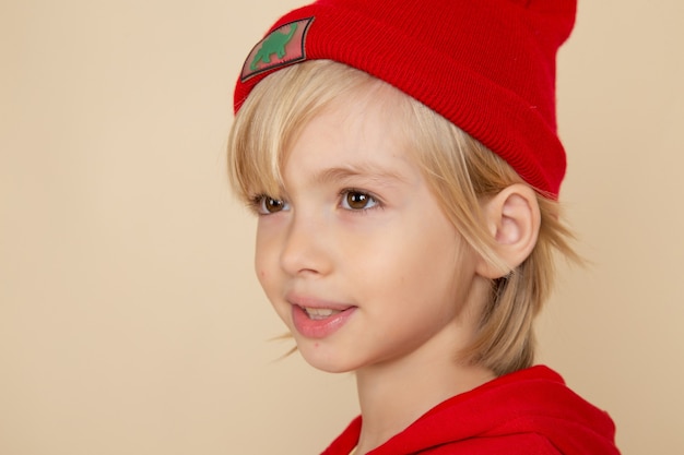 Vista frontal niño lindo en camisa roja y gorra en la pared blanca