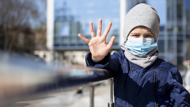 Vista frontal del niño mostrando la mano mientras usa una máscara médica afuera