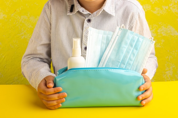 Vista frontal niño pequeño con caja de bolígrafo azul con spray y máscaras sobre superficie amarilla