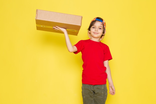 Una vista frontal del niño pequeño en la camiseta roja colorida gorra y pantalón de color caqui con caja en el fondo amarillo