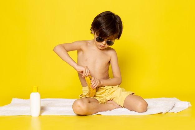 Vista frontal del niño pequeño en gafas de sol pelando plátano en el amarillo