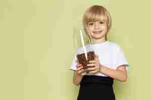 Foto gratuita una vista frontal niño rubio sonriente en camiseta blanca con vaso con semillas de café marrón en el piso de color piedra