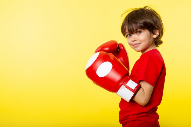 Una vista frontal niño sonriente en camiseta roja y guantes rojos de boxeo en la pared amarilla