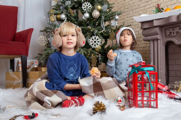Vista frontal de los niños lindos sentados alrededor del árbol de navidad y presenta en su casa color año nuevo infancia navidad