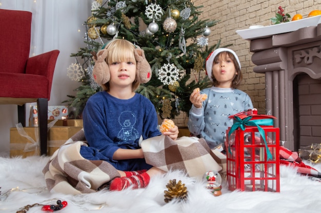 Vista frontal de los niños lindos sentados alrededor del árbol de navidad y presenta en su casa color año nuevo infancia navidad