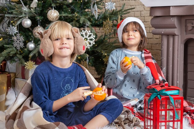 Vista frontal de niños lindos sentados alrededor del árbol de Navidad y regalos en su casa comiendo mandarina kid