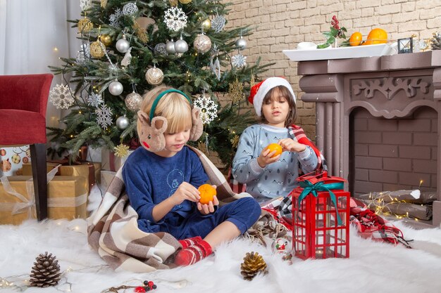 Vista frontal de niños lindos sentados alrededor del árbol de Navidad y regalos en su casa comiendo mandarina niño color foto de año nuevo infancia navidad