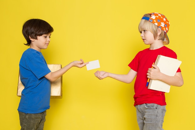 Una vista frontal de niños pequeños en camisetas rojas y azules, gorra de colores y jeans grises con caja en el fondo amarillo