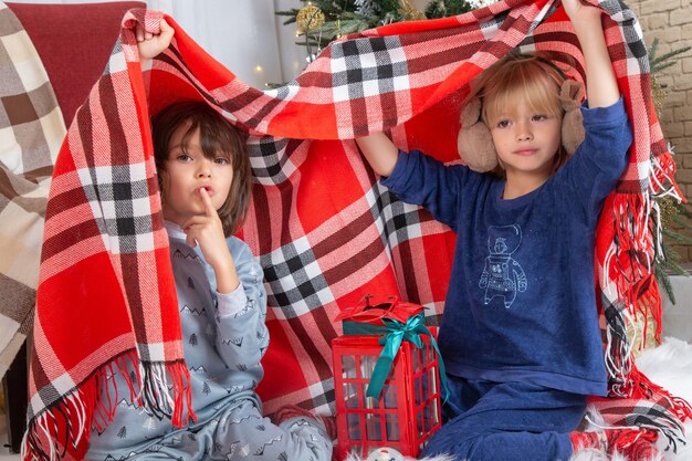 Vista frontal de los niños pequeños lindos que se esconden con la hoja a cuadros alrededor del árbol de Navidad y regalos