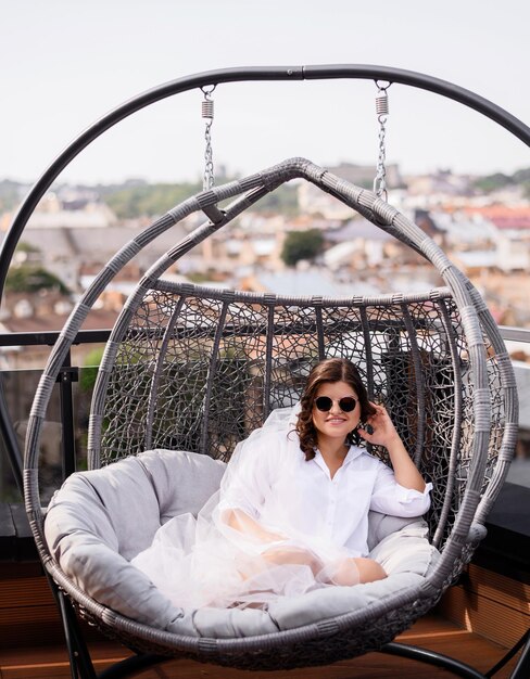 Vista frontal de una novia alegre vestida con un elegante velo largo y gafas de sol sonriendo y mirando a la cámara mientras se sienta en una silla giratoria en la terraza al fondo de una ciudad increíble