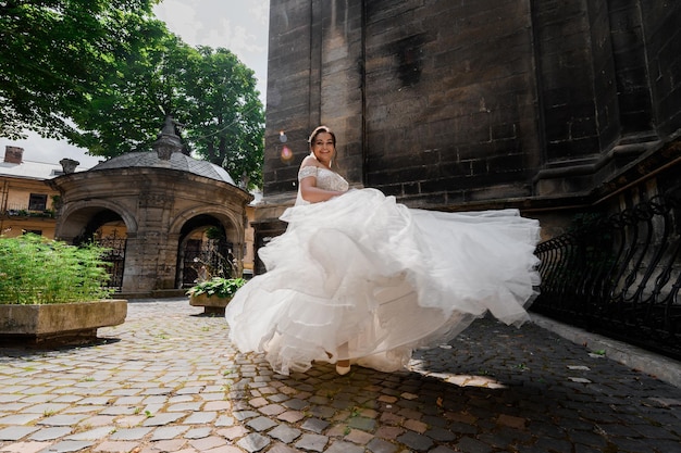 Vista frontal de una novia atractiva con un vestido lujoso que agita su falda sonriendo y mirando la cámara mientras camina cerca de edificios antiguos durante el día soleado de la boda xA