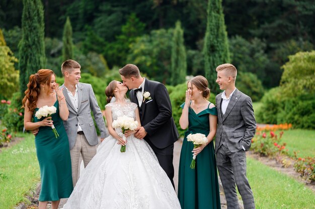 Vista frontal de la novia y el novio felices en ropa de boda de pie entre sus amigos y besándose