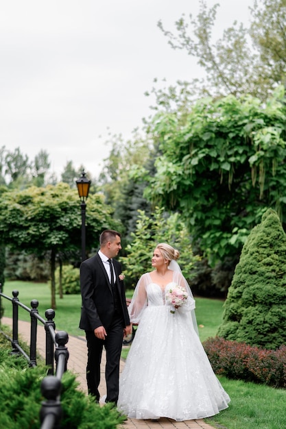 Vista frontal de la novia y el novio felices con ropa elegante caminando por la carretera a través de un parque increíble con hermosas plantas y faroles antiguos sonriendo y mirándose el uno al otro durante el día de la boda