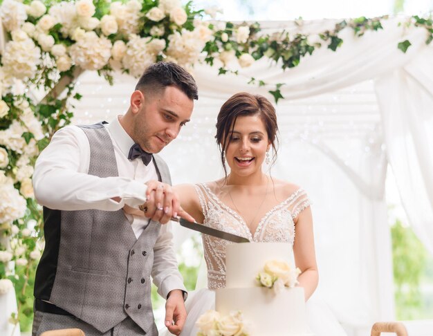 Vista frontal de la novia y el novio felices sosteniendo juntos un cuchillo concentrándose en cortar el pastel de bodas mientras están de pie en el fondo