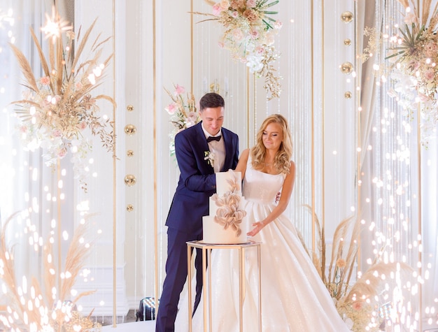 Vista frontal de la novia y el novio de pie en el escenario brillante con decoración cortando el pastel de bodas juntos