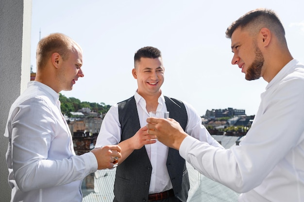 Foto gratuita vista frontal del novio con amigos padrinos de pie sosteniendo copas con paisaje de la ciudad en el fondo celebrando la fiesta del día de la boda hombres amigos saludan a guapo prometido en traje