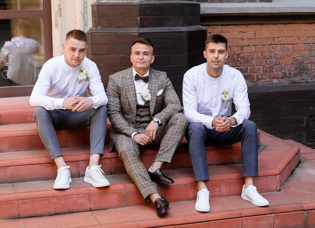 Vista frontal del novio confiado en traje de moda y corbata de moño sentado con amigos en la escalera en el fondo de un edificio antiguo mirando y posando a la cámara en el día de la boda