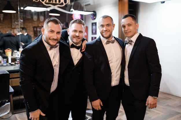 Vista frontal del novio y sus amigos vestidos con trajes festivos negros abrazándose mientras posan y miran la cámara en la barbería el día de la boda