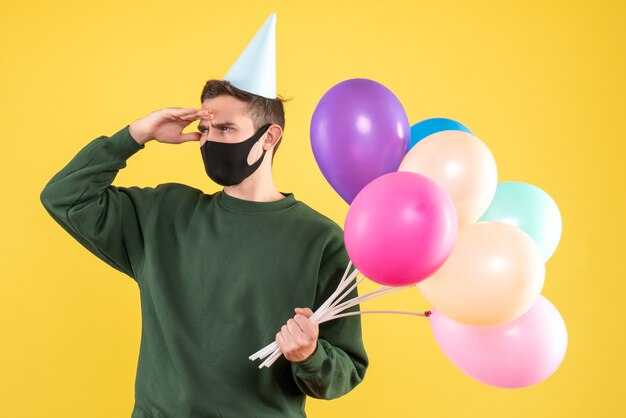 Vista frontal observando a joven con gorro de fiesta y globos de colores de pie sobre fondo amarillo