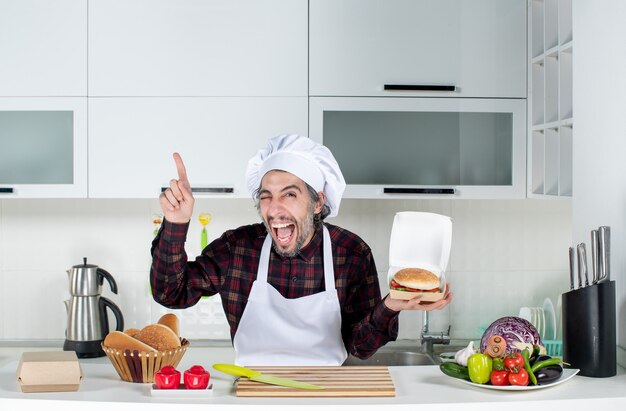 Vista frontal del ojo parpadeante del chef masculino sosteniendo una hamburguesa apuntando al techo de la cocina