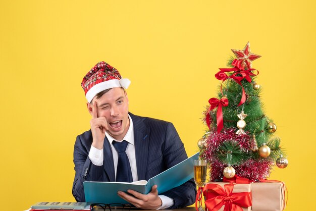 Vista frontal del ojo parpadeante del hombre de negocios sentado en la mesa cerca del árbol de Navidad y presenta en la pared amarilla