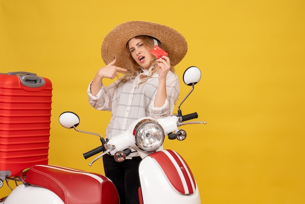 Vista frontal de la orgullosa y ambiciosa mujer joven con sombrero recogiendo su equipaje sentado en una motocicleta y sosteniendo una tarjeta bancaria