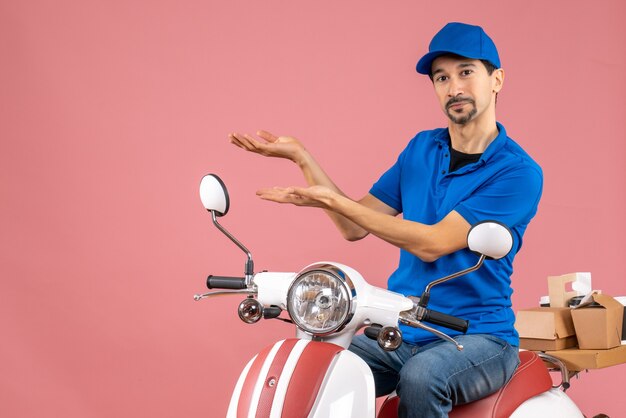 Vista frontal del orgulloso mensajero con sombrero sentado en scooter sobre fondo de melocotón pastel