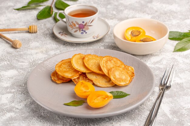 Vista frontal de panqueques dulces dentro de la placa con albaricoques y té en el escritorio gris comida de panqueques postre dulce fruta